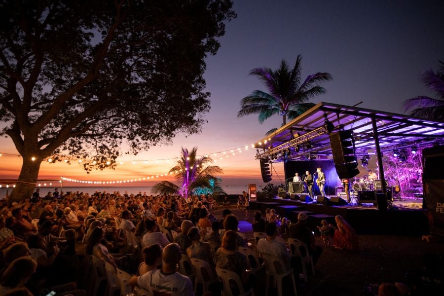 Darwin Festival's closing weekend at the Ski Club includes an array of live bands in 2026, including TISM. The photo depicts a large seated audience watching a band play on an outdoor stage; a tropical sunset over the ocean is visible behind them.