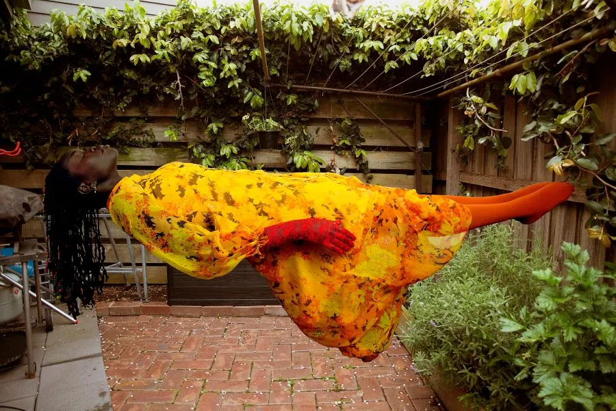 Atong Atem, Our neighbour was a witch (2022) from the Surat series (2022). National Gallery of Victoria, Melbourne. Photo: Atong Atem. A Black woman floats in the air in an inner city suburban courtyard; her beaded hair dangles down towards the brick paving; she wears a bright yellow dress, long red gloves and orange stockings without shoes. A clothesline stretches incongruously above her, and plants grow in the garden and hang from the wooden fences visible beside and behind her.