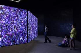 Khaled Sabsabi's artwork conference of one's self, 2026 installed at the Australia Pavilion at the 2026 Venice Biennale. A wall-size video screen projection showing abstract purple patterns. Three viewers stand in a darkened space around it.