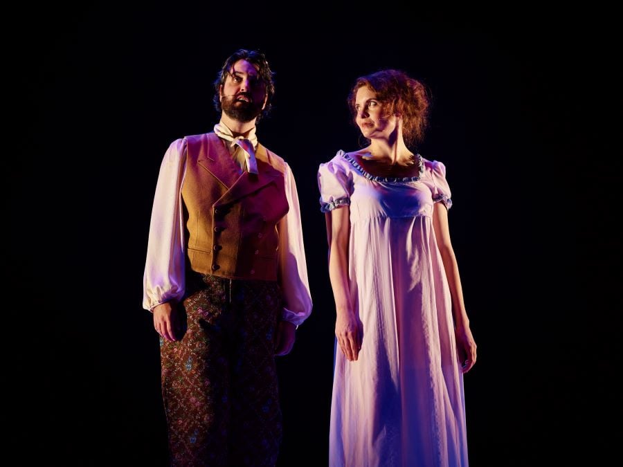 L-R: James Jackson as Mr Darcy and Lauren Swain as Elizabeth Bennet in Bloomshed's Pride and Prejudice at Malthouse Theatre, 2026. Jackson, fair-skinned, dark haired and bearded, looks out at the audience and speaks. Swain, fair-skinned with red hair and wearing a short-sleeved pink Regency-style dress, gazes at him with warmth and humour.