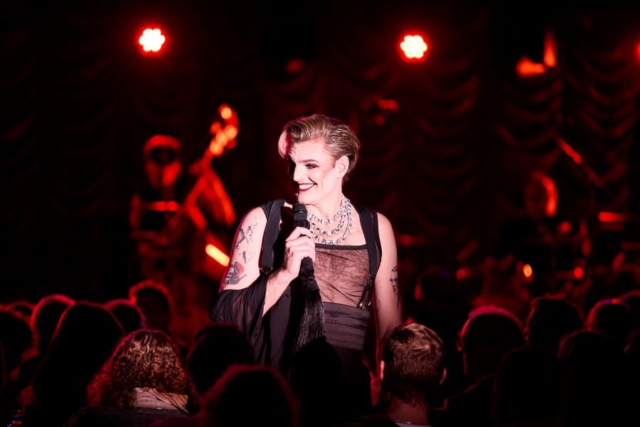 Reuben Kaye, the Artistic Director of the 2026 Adelaide Cabaret Festival, photographed mid-performance amidst his audience. Kaye is a fair-skinned man wearing a transparent, sleeveless mesh vest, thick black braces and a black cummerbund around his waist. A musician is visible on stage behind him, although Kaye is offstage in the midst of his seated audience, whose backs are to the camera. 