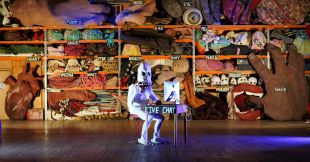 Iconic Victorian company Snuff Puppets has had its annual funding of $112,000 cut by Creative Victoria. Pictured is the Snuff Puppets' popular character Skullie, 2020. A person dressed in a skeleton costume sits at a wooden desk in an old hall. Racks of puppet parts and costume are stored behind it.