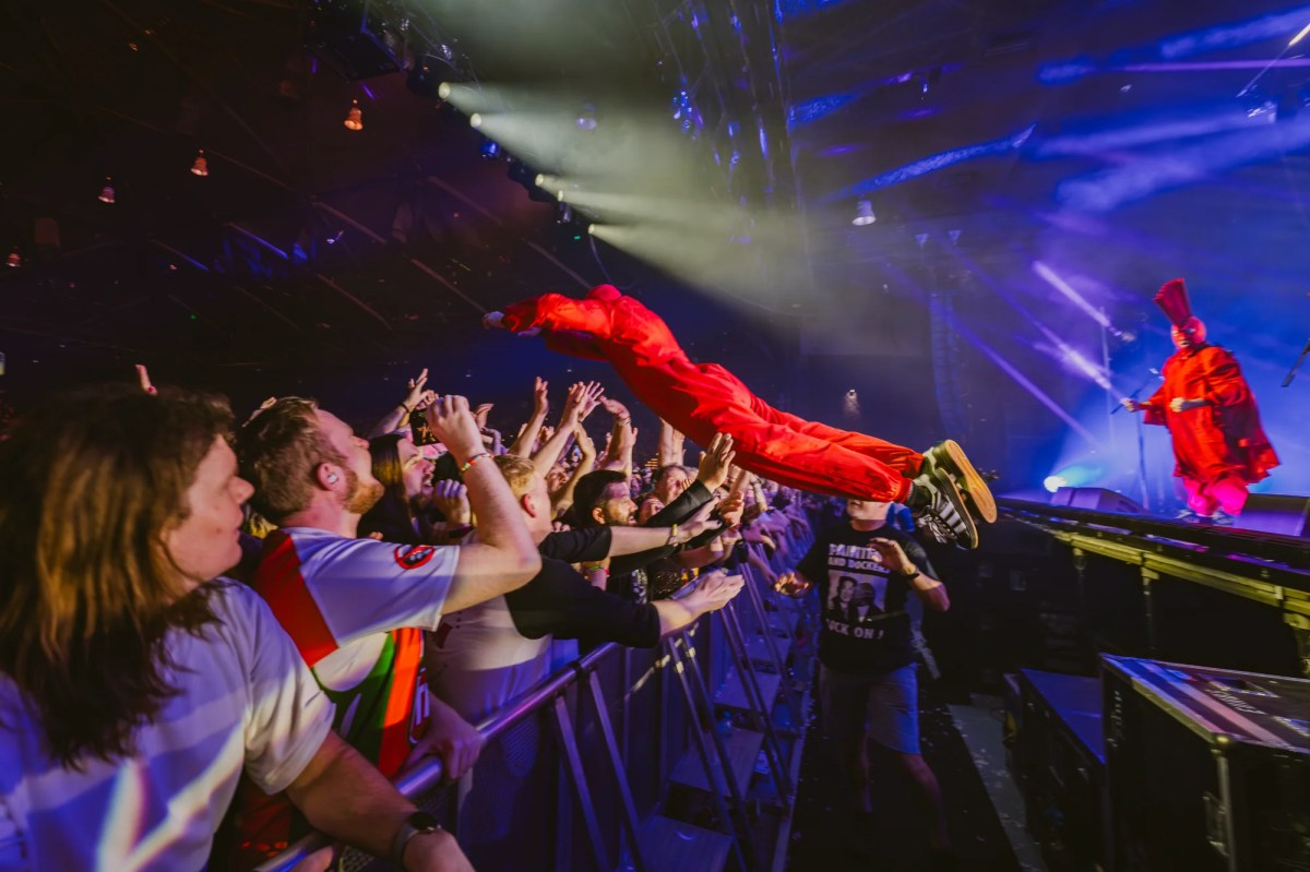 Cult band TISM return to the NT for the 2026 Darwin Festival. TISM last played Darwin in 1996, their only gig there to date. The photo shows one member of TISM, dressed in bright red overalls and a matching balaclava, launching himself off the stage towards the crowd; fans stand ready to catch him, arms upraised, behind the crowd barrier. Another TISM member, also dressed in red overalls but wearing a red balaclava and red, rooster-like crest, watches on from the stage.