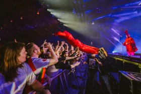 Cult band TISM return to the NT for the 2026 Darwin Festival. TISM last played Darwin in 1996, their only gig there to date. The photo shows one member of TISM, dressed in bright red overalls and a matching balaclava, launching himself off the stage towards the crowd; fans stand ready to catch him, arms upraised, behind the crowd barrier. Another TISM member, also dressed in red overalls but wearing a red balaclava and red, rooster-like crest, watches on from the stage.