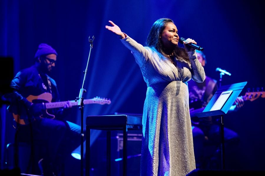 Ursula Yovich Sings Nina Simone at Adelaide Cabaret Festival 2026. Yovich, a Burarra woman, wears a white dress and stands with her right arm raised; in her left hand she holds a microphone close to her mouth. Musicians are visible on stage behind her. 