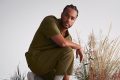 Poetry Month 2026 international guest Jericho Brown (US). The photo shows a Black American man standing in a half-crouched position with tall grass growing behind him.