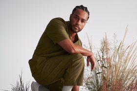 Poetry Month 2026 international guest Jericho Brown (US). The photo shows a Black American man standing in a half-crouched position with tall grass growing behind him.