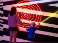 A mother and child, photographed from behind, admire Brook Andrew's neon light and painted wall artwork Loop. A model of how the world operates (detail), 2008, installation view MCA Collection: Artists in Focus, Museum of Contemporary Art Australia, 2026. Credit: Maja Baska. arts participation