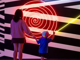 A mother and child, photographed from behind, admire Brook Andrew's neon light and painted wall artwork Loop. A model of how the world operates (detail), 2008, installation view MCA Collection: Artists in Focus, Museum of Contemporary Art Australia, 2026. Credit: Maja Baska. arts participation