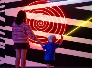 A mother and child, photographed from behind, admire Brook Andrew's neon light and painted wall artwork Loop. A model of how the world operates (detail), 2008, installation view MCA Collection: Artists in Focus, Museum of Contemporary Art Australia, 2026. Credit: Maja Baska. arts participation