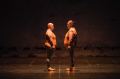 A scene from Back to Back Theatre's 2008 work, Food Court. Two plump figures, identically dressed in sleeveless orange vinyl tops, grey leggings and orange sneakers, face each other on a bare stage.