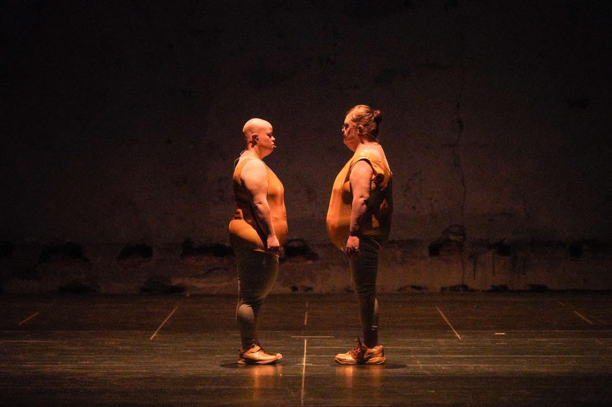 A scene from Back to Back Theatre's 2008 work, Food Court. Two plump figures, identically dressed in sleeveless orange vinyl tops, grey leggings and orange sneakers, face each other on a bare stage.