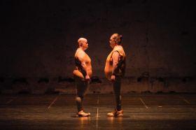 A scene from Back to Back Theatre's 2008 work, Food Court. Two plump figures, identically dressed in sleeveless orange vinyl tops, grey leggings and orange sneakers, face each other on a bare stage.