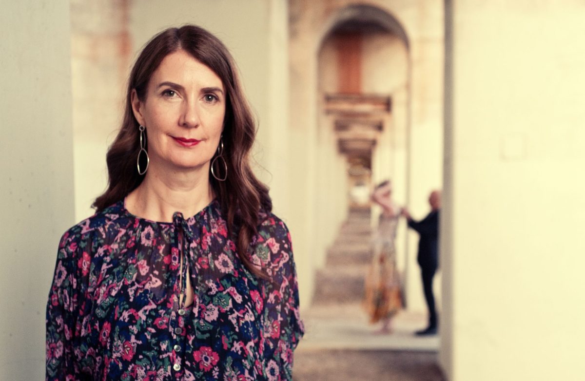 Anna Goldsworthy's Quarterly Essay explores AI, the future and what it means to be human as technology rapidly learns to mirror us. The photo shows Goldsworthy, a fair skinned woman with long dark hair and wearing a pink, purple and green pattened dress, standing in a colonnade.