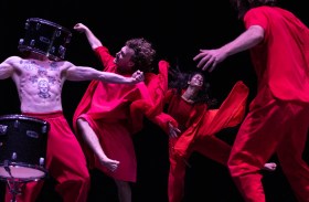 contemporary dance: a photo of four dancers, three of whom are dressed in a bright red costume moving vigorously on stage against a black background. One of the four, a male dancer, is bare-chested and his head is covered with a drum (musical instrument). There is another drum on stage pictured at the of the photo.