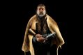 Noongar composer Aaron Wyatt in a promotional image for Decolonising the Bells, his collaboration with Speak Percussion, which premieres at Kindred People in September. An Aboriginal man with long hair, a short grey beard and glasses, faces the camera. We wears a kangaroo-skin cloak and holds a handbell in one hand and a painted stick in the other.