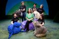 Eva Di Cesare, an older woman with dark, greying, shoulder length hair, surrounded by performers and characters from Monkey Baa's many productions. Eva wears a green shirt and black pants, and sits with her legs crossed. The people around her wear black 'Monkey Baa' t-shirts and hold an array of puppets including an emu, a wombat and a blue sheep.