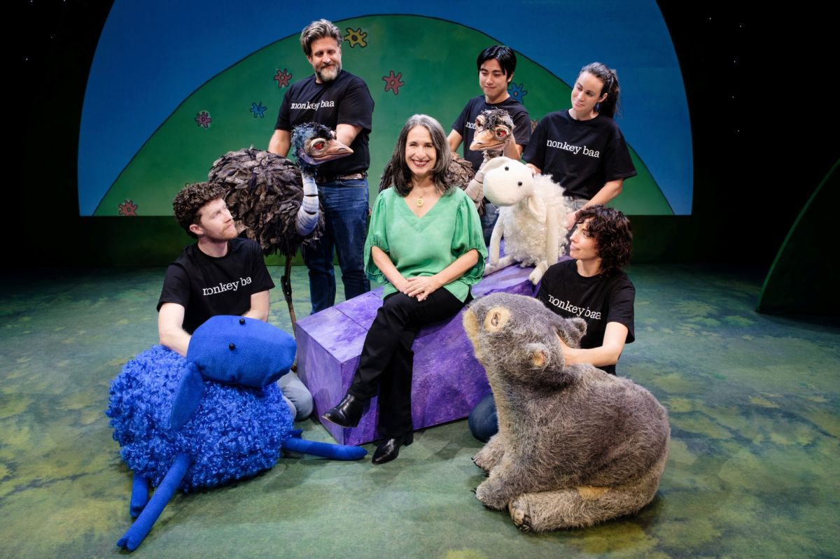 Eva Di Cesare, an older woman with dark, greying, shoulder length hair, surrounded by performers and characters from Monkey Baa's many productions. Eva wears a green shirt and black pants, and sits with her legs crossed. The people around her wear black 'Monkey Baa' t-shirts and hold an array of puppets including an emu, a wombat and a blue sheep.