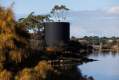 In Absence Mona Yhonnie Scarce: a photo of a large ciccular ashen-coloured wooden tower installed amongst trees on the edge of a river which is Berriedale Bay in Nipaluna/ Hobart.