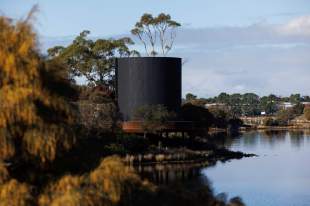 In Absence Mona Yhonnie Scarce: a photo of a large ciccular ashen-coloured wooden tower installed amongst trees on the edge of a river which is Berriedale Bay in Nipaluna/ Hobart.