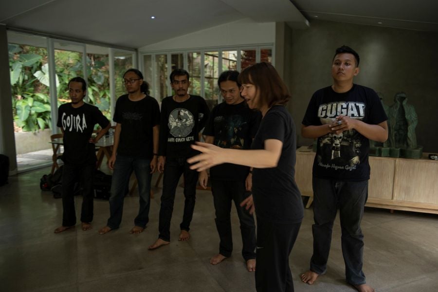 Lucy Guerin AO in the studio for LGI's 2020 production Metal. Guerin, a slender, middle-aged woman with fair skin and shoulder length dark hair, gestures emphatically. Five Indonesian men from Ensemble Tikoro, a heavy metal choir, stand behind her; they are wearing black band t-shirts, black jeans and are barefoot.