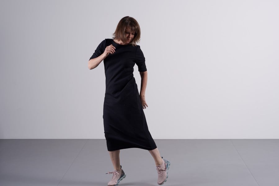 A photograph of Lucy Guerin dancing, her right arm raised, her left arm extended by her side. Her brown, shoulder-length hair swings; she is fair skinned and wearing a black dress.