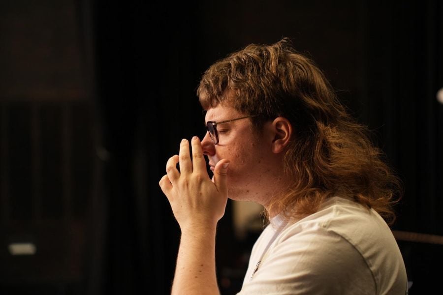 Back to Back Theatres in-development work Neanderthal. The photo depicts a young man with a serious mullet photographed in profile. He wears glasses and raises his left hand towards his face.