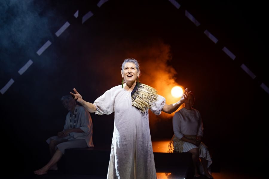 A scene from the Sydney Opera House season of UPU. A performer dressed in white gestures dramatically as she declaims a poem. Two more poet-performers sit behind her, half seen. 