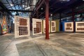 Khaled Sabsabi Between Suns: Installation view of an exhibition showing a series of large free-hanging canvases painted with brown graphic patterns in an industrial warehouse gallery space.
