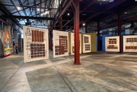Khaled Sabsabi Between Suns: Installation view of an exhibition showing a series of large free-hanging canvases painted with brown graphic patterns in an industrial warehouse gallery space.