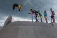 Snake Sessions performance in public space: a photo of four young male people standing at the top of a skate park ramp watching another young male person do a somersault in the air.