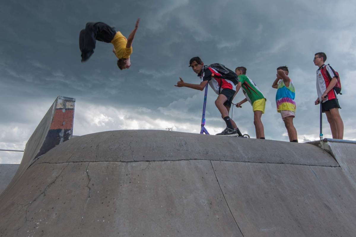 Snake Sessions performance in public space: a photo of four young male people standing at the top of a skate park ramp watching another young male person do a somersault in the air.