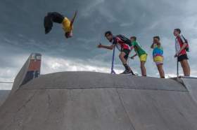 Snake Sessions performance in public space: a photo of four young male people standing at the top of a skate park ramp watching another young male person do a somersault in the air.
