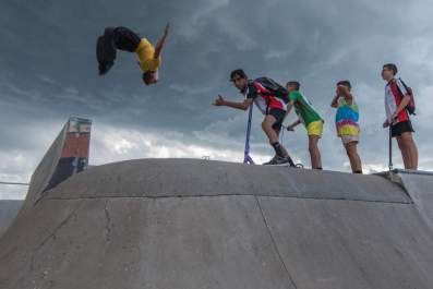 Snake Sessions performance in public space: a photo of four young male people standing at the top of a skate park ramp watching another young male person do a somersault in the air.